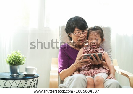 portrait photo of elderly or old Asian retirement woman smiling and watching on smartphone while siting with her granddaughter on armchair in living room. technology, communication and people concept