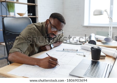 Afro-American architect working in office with blueprints.Engineer inspect architectural plan, sketching a construction project. Portrait of black handsome man sitting at workplace. Business concept. Royalty-Free Stock Photo #1806903694
