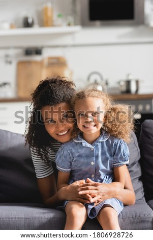 young african american mother embracing daughter while sitting on sofa and looking at camera