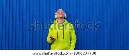 close up and portrait of teenager young man listening music and having fun with his phone and wearing headphones - blue background wall - cheerful and happy millennial hearing music ad vibing Royalty-Free Stock Photo #1806937738
