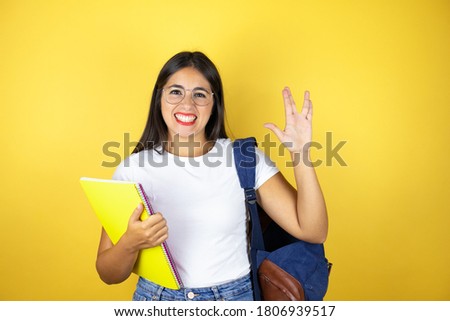 Young beautiful student woman wearing backpack doing hand symbol holding notebook over isolated yellow background