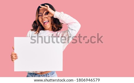 Young beautiful mixed race woman holding cardboard banner with blank space smiling happy doing ok sign with hand on eye looking through fingers 