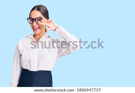 Beautiful brunette young woman wearing professional waitress apron doing peace symbol with fingers over face, smiling cheerful showing victory 