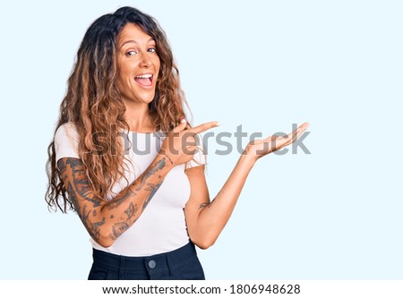 Young hispanic woman with tattoo wearing casual white tshirt amazed and smiling to the camera while presenting with hand and pointing with finger. 