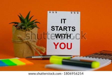White note with inscription 'it starts with you' on beautiful orange background, colored paper, metalic pen, yellow marker,  flowerpot with a plant and calculator. Business concept.