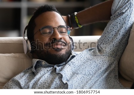 Close up satisfied African American man wearing headphones enjoying favorite music with closed eyes, daydreaming or sleeping, positive young male wearing glasses lying relaxing on cozy couch Royalty-Free Stock Photo #1806957754
