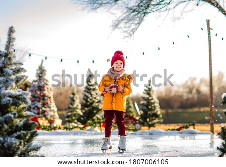 Young cute boy in orange winter jacket and red hat holding Santa's cup of cacao with marshmallows standing on a fake ice rink among Christmas trees.