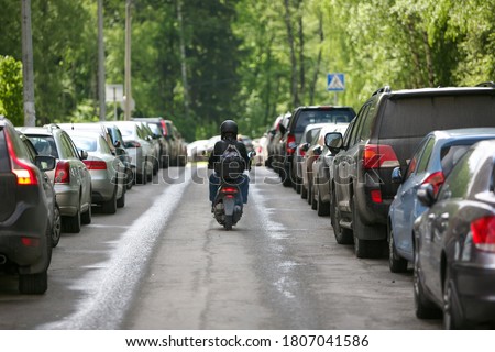 A man on a scooter drives off into the distance among parked cars against a background of green trees. Travel by Moto. Royalty-Free Stock Photo #1807041586