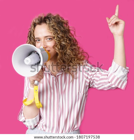Young woman speaks into a megaphone and raises her finger up. Isolated pink background. People sincere emotions lifestyle concept.