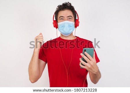 Positive Young caucasian man with short hair wearing medical mask standing over isolated white background holds modern cell phone connected to headphones, clenches fist from good emotions, exclaims 