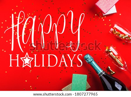 Wishing happy holidays text. Champagne bottle, gifts and flutes with golden streamer on classic festive red background. Celebrating new year, christmas concept festive flat lay.