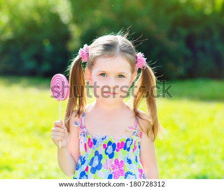 Adorable little girl with two blond ponytails holding white and pink lollipop in her hand, outdoor summer portrait