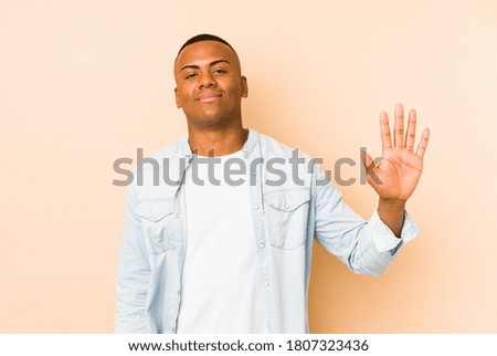 Young latin man isolated on beige background smiling cheerful showing number five with fingers.