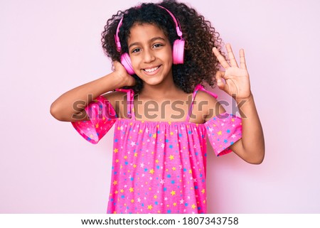 African american child with curly hair listening to music using headphones doing ok sign with fingers, smiling friendly gesturing excellent symbol 