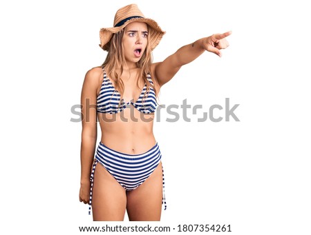 Young beautiful blonde girl wearing bikini and hat pointing with finger surprised ahead, open mouth amazed expression, something on the front 