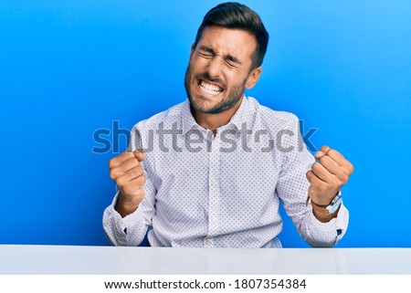 Handsome hispanic man wearing business clothes sitting on the table very happy and excited doing winner gesture with arms raised, smiling and screaming for success. celebration concept. 