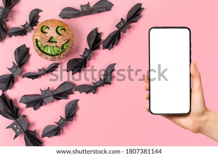 A females hand holds a smartphone. In the left corner is a pumpkin lantern and flock of bats made of paper. Purple background. Mock up. The concept of Halloween and technologies