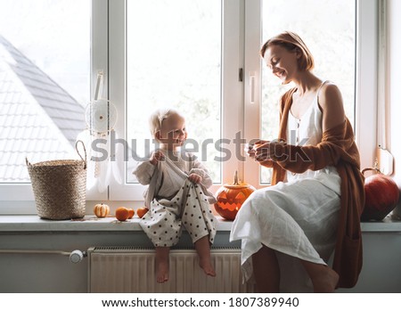 Minimalist style family with child preparing for Halloween. Happy mother and her little daughter sitting near window with carving pumpkin head jack lantern and halloween decoration at home.