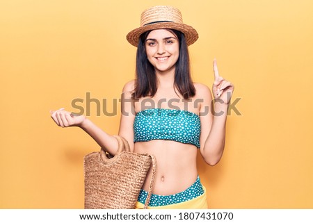 Young beautiful girl wearing bikini and hat holding bag smiling with an idea or question pointing finger with happy face, number one 