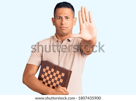 Young handsome latin man holding chess with open hand doing stop sign with serious and confident expression, defense gesture 
