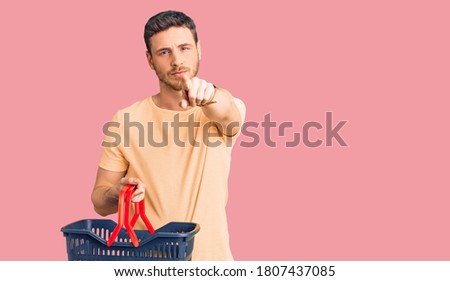 Handsome young man with bear holding supermarket shopping basket pointing with finger to the camera and to you, confident gesture looking serious 