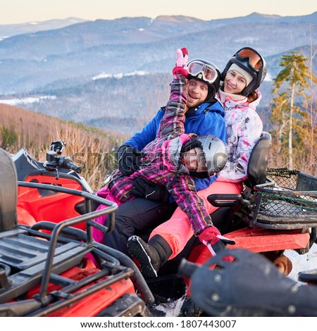 Happy family of three, wearing colorful ski suits, spending great winter holiday, hugging together in snowy mountains on red quad bike, side view, ridge of mountains on background.