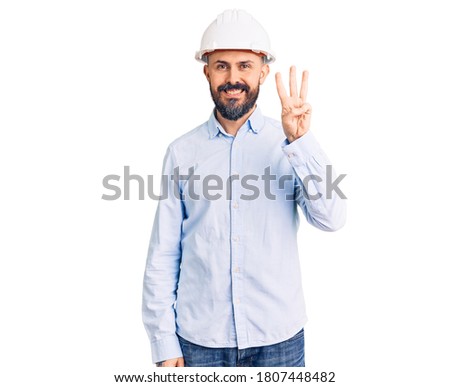 Young handsome man wearing architect hardhat showing and pointing up with fingers number three while smiling confident and happy. 