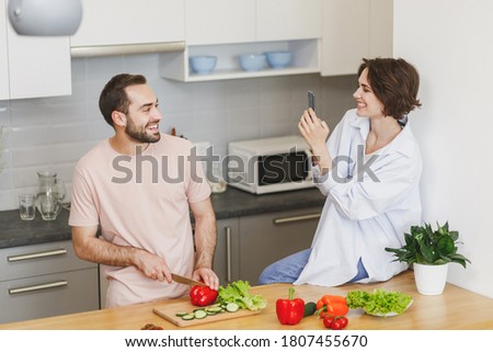 Funny calm happy lovely young family couple woman and man in casual t-shirts clothes, sitting on table, taking pictures on mobile phone preparing vegetable salad cooking food in light kitchen at home.
