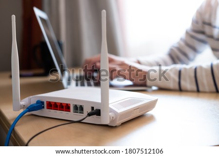 Selective focus at router. Internet router on working table with blurred man connect the cable at the background. Fast and high speed internet connection from fiber line with LAN cable connection. Royalty-Free Stock Photo #1807512106