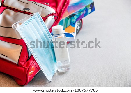 Detail of a kid's red backpack with a blue disposable mask and hand sanitizer set ready for back to school