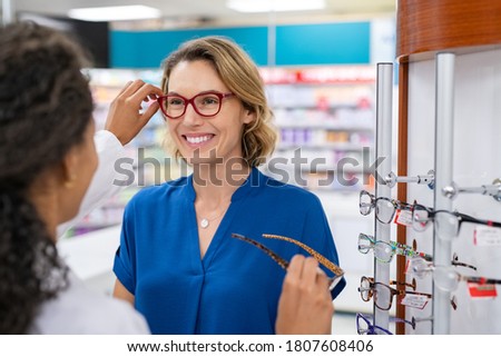 Mid adult woman in optic store buying new glasses. Happy smiling woman trying new eyeglasses with optician in pharmacy. Happy woman trying new spectacles frame in optic store. Royalty-Free Stock Photo #1807608406