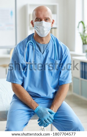 Vertical medium long portriat of unrecognizable doctor wearing blue uniform, latex gloves and mask on face sitting relaxed looking at camera