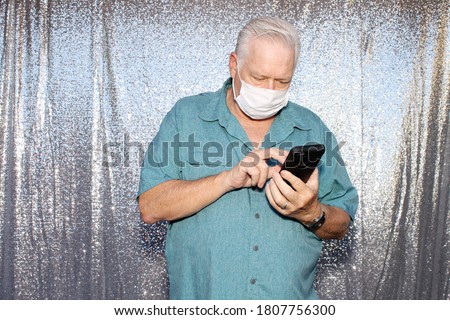 Photo Booth. A  Caucasian Man wears a Anti-Covid 19 Face Mask sends text messages from his phone as he poses for photos in a Photo Booth with a silver sequin background.  
