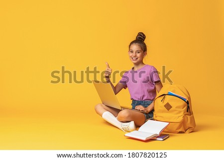happy schoolgirl with backpack, laptop and books is sitting on yellow background in the Studio. The child shows, a sign of approval, a finger up. Back to school. copy space.