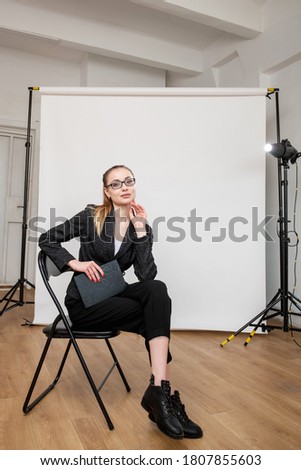 Shooting backstage. Professional photography. Woman in black suit posing sitting in modern studio with light equipment white backdrop. Creative work. Business people lifestyle.