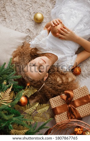 A happy little girl lies under a Christmas tree with gifts on the carpet. The view from the top