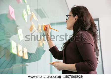 Confident middle-aged businesswoman writing on sticker with pencil and brainstorming. Colorful memo notes stuck on glass wall in conference room. Business, planning and management concept Royalty-Free Stock Photo #1807868929
