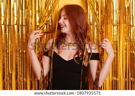 Pretty female clubber being photographed among golden tinsel, looking smiling aside, receives congratulation with birthday, laughs positively, wearing beautiful black dress.