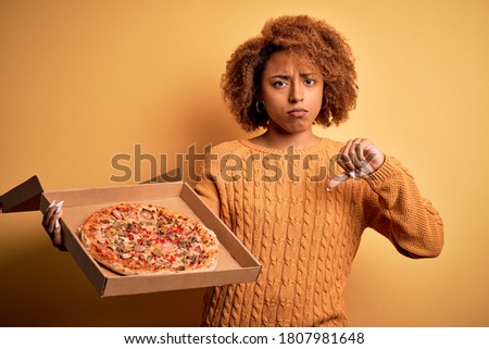 Young African American afro woman with curly hair holding delivery box with Italian fresh pizza with angry face, negative sign showing dislike with thumbs down, rejection concept