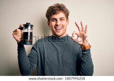 Young blond man with curly hair making coffee using coffemaker over white background doing ok sign with fingers, excellent symbol