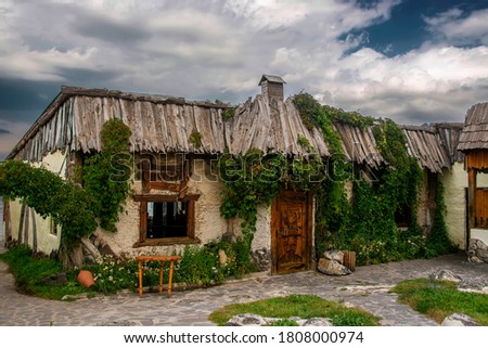 Old dreamlike house with wooden roof, door, window and clay walls covered with green beautiful plants under the cloudy blue dramatic sky. Translation: "lagoon" written in Armenian