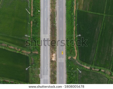 Aerial photograph of a yellow car running on a paved road that cuts through the green fields.