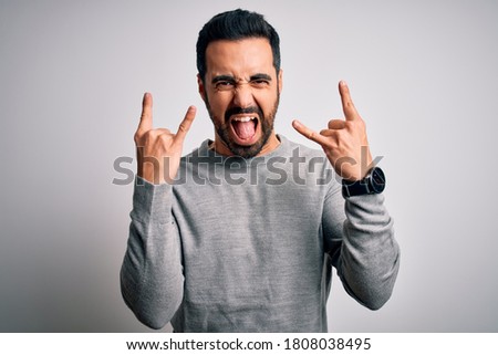 Young handsome man with beard wearing casual sweater standing over white background shouting with crazy expression doing rock symbol with hands up. Music star. Heavy concept.