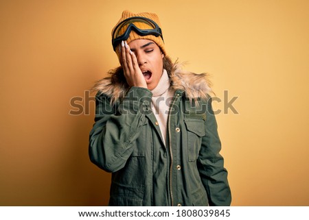 Young african american afro skier girl wearing snow sportswear and ski goggles Yawning tired covering half face, eye and mouth with hand. Face hurts in pain.