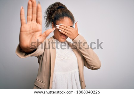 Beautiful african american businesswoman wearing jacket and glasses over white background covering eyes with hands and doing stop gesture with sad and fear expression. Embarrassed and negative concept