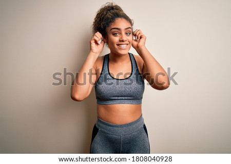 Young african american sportswoman doing sport wearing sportswear over white background Smiling pulling ears with fingers, funny gesture. Audition problem