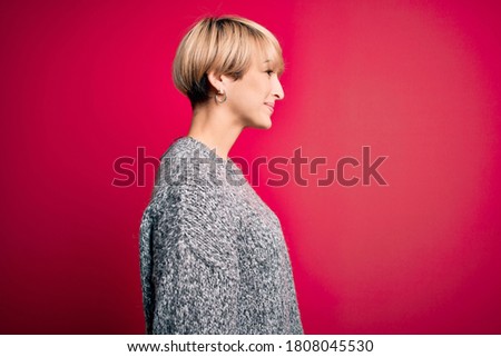 Young blonde woman with modern short hair wearing casual sweater over pink background looking to side, relax profile pose with natural face and confident smile.