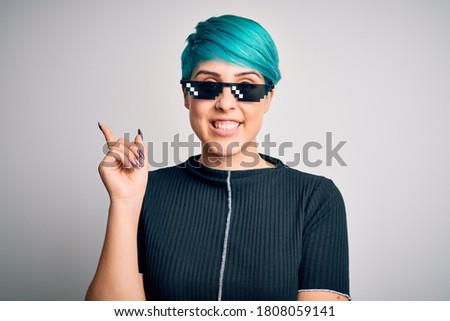 Young woman with blue fashion hair wearing thug life sunglasses over white background with a big smile on face, pointing with hand and finger to the side looking at the camera.
