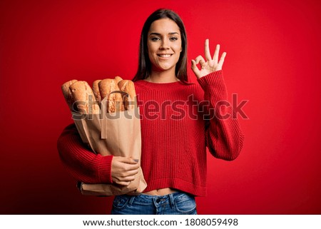 Young beautiful brunette woman holding paper bag with bread over red background doing ok sign with fingers, excellent symbol