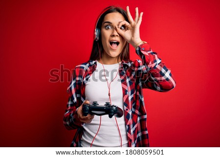 Young beautiful brunette gamer woman playing video game using joystick and headphones with happy face smiling doing ok sign with hand on eye looking through fingers
